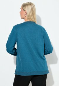 Maglia a maniche lunghe color verde acqua con scollatura rotonda, vestibilità comoda e orlo a coste. Tessuto morbido con una texture liscia; indossata sopra pantaloni neri.