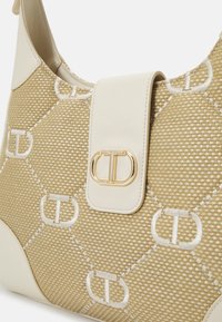 Sac à main beige tissé avec des motifs "TD" brodés en blanc et une sangle en cuir blanc dotée d'une boucle ovale dorée.
