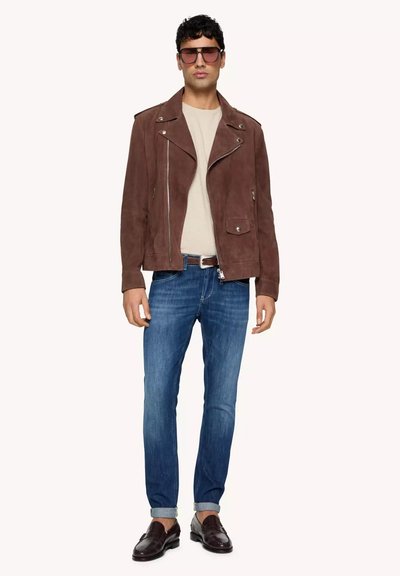 Giacca motociclistica in suede marrone con cerniere argentate, camicia crema, jeans in denim blu e mocassini scuri. Gli accessori includono occhiali da sole grandi.