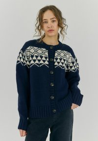 Námornícky modrý pletený kardigán s bielym geometrickým vzorom fair isle, rebrovanými manžetami a lemom, a čiernymi gombíkmi na prednej strane.