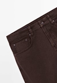 Jean en denim brun foncé, visible de la taille jusqu'au milieu de la cuisse, avec bouton devant, passants de ceinture et une petite poche à monnaie sur fond blanc.