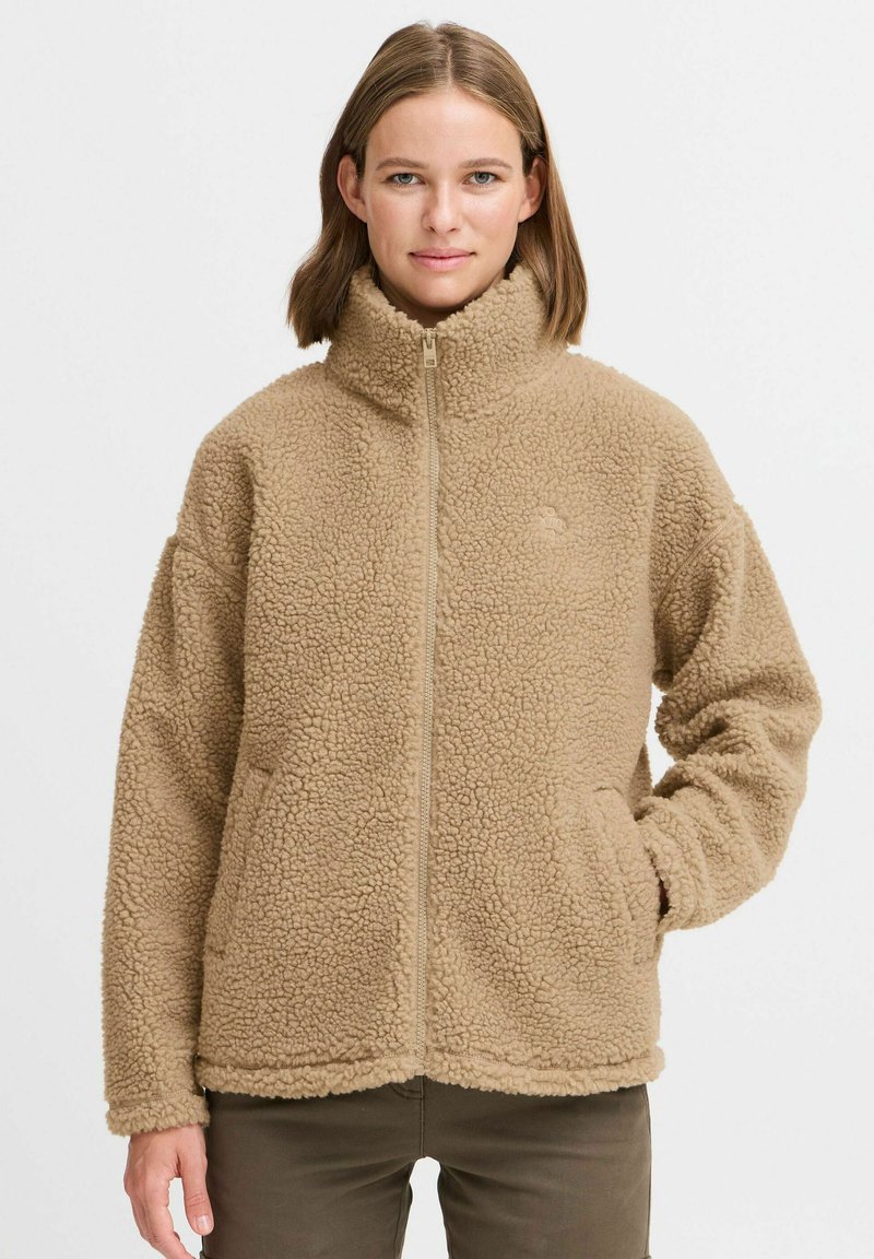 Veste en polaire beige avec un col montant, fermeture éclair et poches latérales. La texture est pelucheuse, avec un tissu doux et confortable sur l'ensemble.