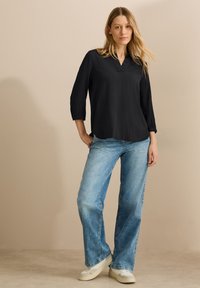 Schwarze, strukturierte Bluse mit V-Ausschnitt und Dreiviertelärmeln, kombiniert mit hellblauen, weitem Jeans und weißen Sneakers.