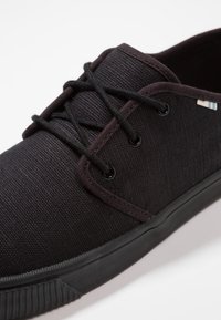 Mustat kangaskengät, joissa on teksturoitu pinta, pyöreä kärki, mustat nauhat ja kumipohja. Varoitus pieni raidoitusvarustus kantapään lähellä.