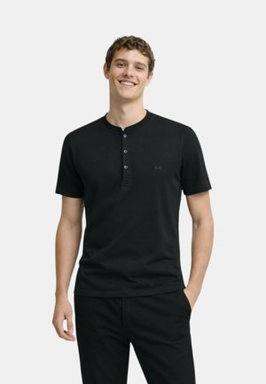 Giovane uomo con indosso una maglietta Henley nera a maniche corte con tre bottoni, in piedi con una mano in tasca, sorridente su uno sfondo semplice.