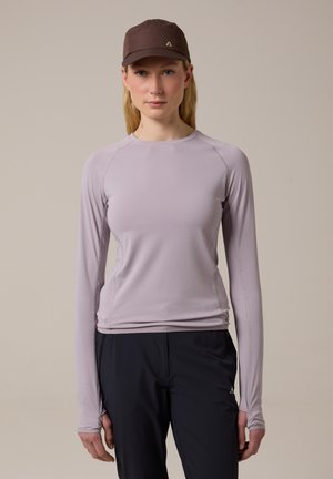 Femme portant une chemise à manches longues violet clair, un pantalon noir et une casquette marron foncé, debout devant un fond neutre.