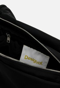 Czarna torba z tkaniny z metalowym zamkiem błyskawicznym. Wnętrze posiada białą metkę z napisem "Desigual" w kolorze żółtym. Gładka faktura.