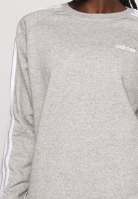 Primer plano de una persona que lleva una sudadera Adidas de color gris claro con tres rayas blancas en las mangas y un pequeño logo de Adidas en blanco en el pecho.