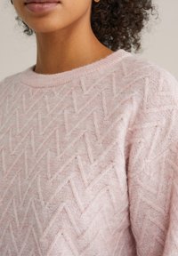 Világos rózsaszín kötött pulóver, texturált cikk-cakk mintával, kerek nyakkivágással és puha, bolyhos anyagból. Közeli kép a részletekről.