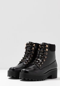 Bottines en cuir verni noir avec une semelle plateforme épaisse, dotées d'œillets en métal doré, de lacets noirs et d'une tige de cheville en tissu noir texturé.