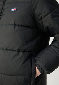 Zwarte gewatteerde jas met zichtbaar Tommy Jeans-logo op de borst en een hand in de zijzak gestoken met elastische manchetten.