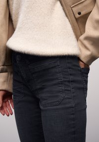 Vaqueros de denim negro con detalles en los bolsillos, combinados con un suéter crema y peludo y una chaqueta beige, mostrando una textura suave y un corte moderno.