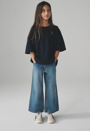 Marinblå oversize t-shirt, vida ljusblå jeans och vita sneakers med marinblå detaljer. Långt hår ramas in kring ansiktet. Enkel, avslappnad outfit.