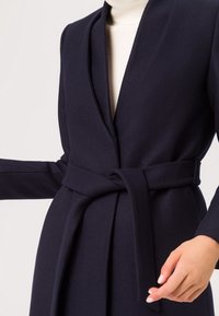 Cappotto avvolgente navy realizzato in tessuto testurizzato, con scollo a V profondo, maniche lunghe e una cintura annodata in vita per esaltare la silhouette.