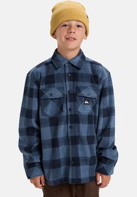 Camicia di flanella a righe blu e navy con bottoni neri e tasche, abbinata a un berretto a maglia giallo. Tessuto caldo e vestibilità comoda.