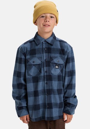 Chemise en flanelle à carreaux bleus et navy avec des boutons et des poches noires, associée à un bonnet en tricot jaune. Tissu confortable et coupe décontractée.