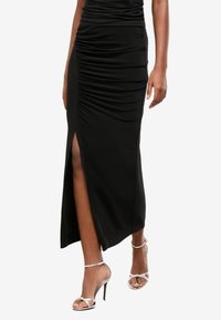 Jupe maxi noire froncée avec fente sur le côté, faite de tissu lisse ; associée à des talons argentés à lanières, arborant une silhouette élancée et un design élégant.