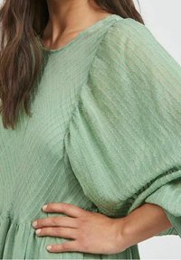 Světlé zelené šaty s texturovaným, žebrovaným designem. Mají nafouknuté rukávy a kulatý výstřih, vyrobené z měkké, splývavé látky.