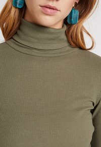 Olivgrünes, geripptes Rollkragenoberteil mit eng anliegendem Design, ergänzt durch große, transparente türkisfarbene Ohrringe mit geometrischer Form.