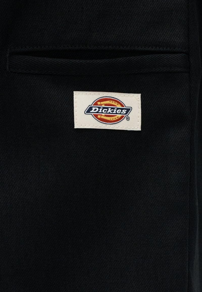 Tessuto nero con una piccola etichetta rettangolare bianca con il logo Dickies in rosso, arancione e blu sopra una tasca orizzontale.