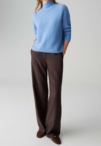 Personne portant un pull en maille bleu clair, un pantalon large marron foncé et des chaussures marron foncé, les mains dans les poches.