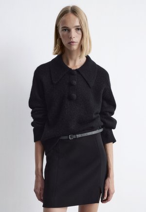 Schwarzer Wollpullover mit einem breiten Kragen und drei Knopfdetails, kombiniert mit einem kurzen schwarzen Rock und einem schmalen grauen Gürtel.