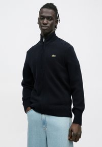 Camisola de malha com fecho éclair azul-marinho, feita de um material de malha texturizada, com gola e punhos canelados, e um pequeno logótipo verde no peito.