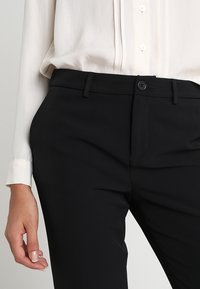 Černé kalhoty na míru s hladkým materiálem, předními kapsami a knoflíkovým zapínáním. Nosené s košilí s knoflíky v světlé barvě.