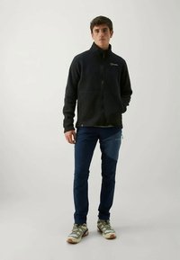 Black fleece jacket with a high collar, zippered front, and logo on chest, paired with blue pants and beige hiking shoes.