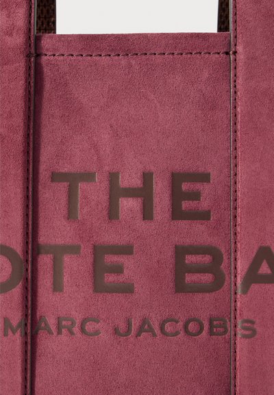 Nærbillede af en lyserød ruskinds taske med mørkebrune præget tekst, der står "THE TOTE BAG MARC JACOBS."