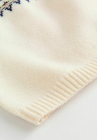 Cremefarvet strikket stof med en tekstureret overflade og ribbede kanter. Subtile mønsterdetaljer i mørke og lyse nuancer langs kanten.