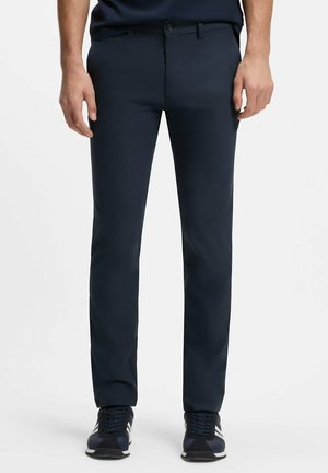 Uomo con pantaloni a vita bassa blu navy aderenti e sneakers abbinate, in piedi su uno sfondo bianco uniforme.