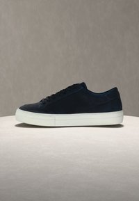 Μαύρο αθλητικό παπούτσι από σουέντ χαμηλού προφίλ με λευκή παχιά σόλα από καουτσούκ και μαύρα κορδόνια, τοποθετημένο σε φωτεινή επιφάνεια με υφή και ουδέτερο φόντο.