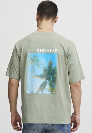 Homme portant un t-shirt vert clair avec un motif de palmier bleu et le mot "ARCHIVE" au dos, accompagné d'un jean en denim bleu.