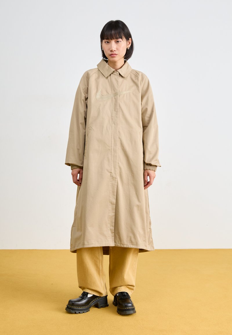 Lang trenchcoat med krage og frontlukking, med et subtilt logo. Kombinert med lysegule bukser og kraftige svarte sko. Ensfarget bakgrunn.