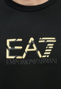 Svart T-skjorte med en fremtredende gull- og svart grafisk logo med teksten "EA7" og "EMPORIO ARMANI". Mykt, teksturert stoff med ribbestrikket hals.
