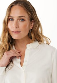 Camicia bianca con colletto, caratterizzata da dettagli sui bottoni ben visibili e una consistenza morbida. Il modello ha capelli mossi e orecchini a cerchio semplici.
