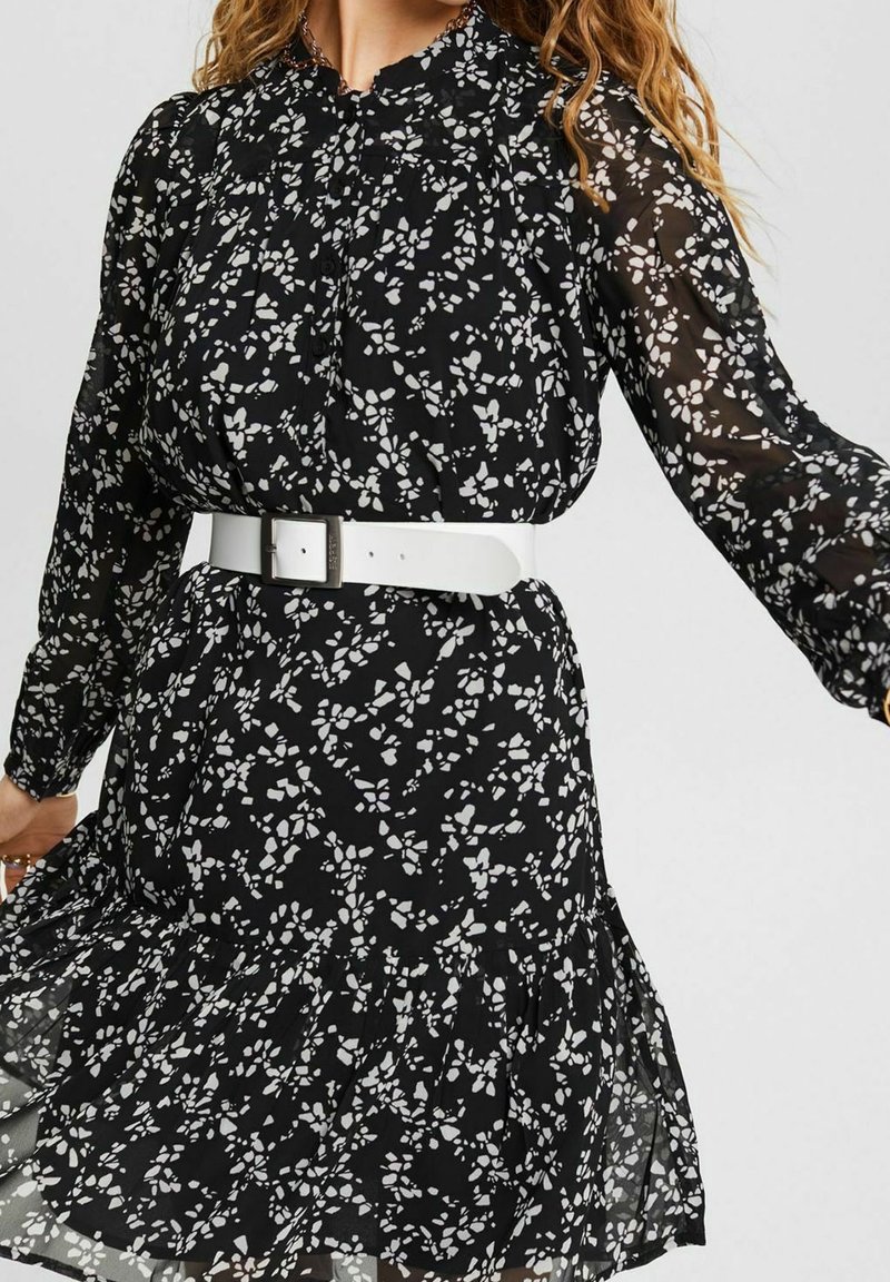 Robe noire à imprimé floral avec tissu transparent, manches longues, ourlet à volants et ceinture blanche à la taille. Met en valeur les détails du motif floral.