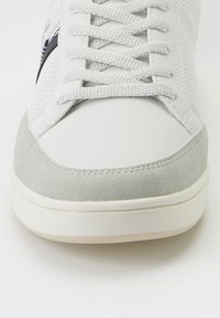 Weißer Sneaker mit grauen Wildlederakzenten, strukturiertem Mesh-Obermaterial, flachen weißen Schnürsenkeln und einer runden Zehenpartie. Besitzt gestickte Details und eine Gummisohle.