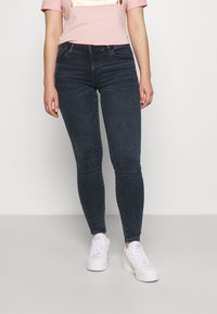 Persona con jeans oscuros ajustados, camiseta de manga corta color rosa claro y zapatillas Adidas blancas, de pie contra un fondo claro y sencillo.