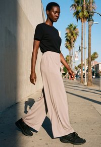 Kvinde går på solrig fortov iført sort skjorte, beige vide bukser og sorte sneakers med palmer og gadelygter i baggrunden.