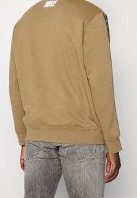 Tan sweatshirt med ribbade muddar och fåll, med en vit Nike-logotyp på baksidan. Matchas med ljusgrå jeans.
