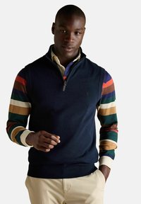 Mężczyzna w granatowym swetrze bez rękawów, noszącym kolorową, w paski koszulę z długim rękawem i beżowe spodnie, pozujący z jedną ręką w kieszeni.