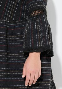 Robe noire avec des rayures verticales multicolores, tissu texturé, manches en cloche et accents en dentelle aux poignets. Main visible, montrant une peau lisse.