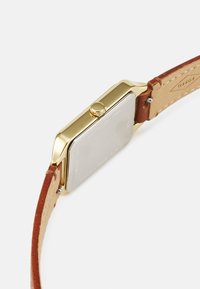 Montre en acier inoxydable doré avec un boîtier carré et un bracelet en cuir marron. Elle présente une couronne et une finition lisse et texturée sur le métal.