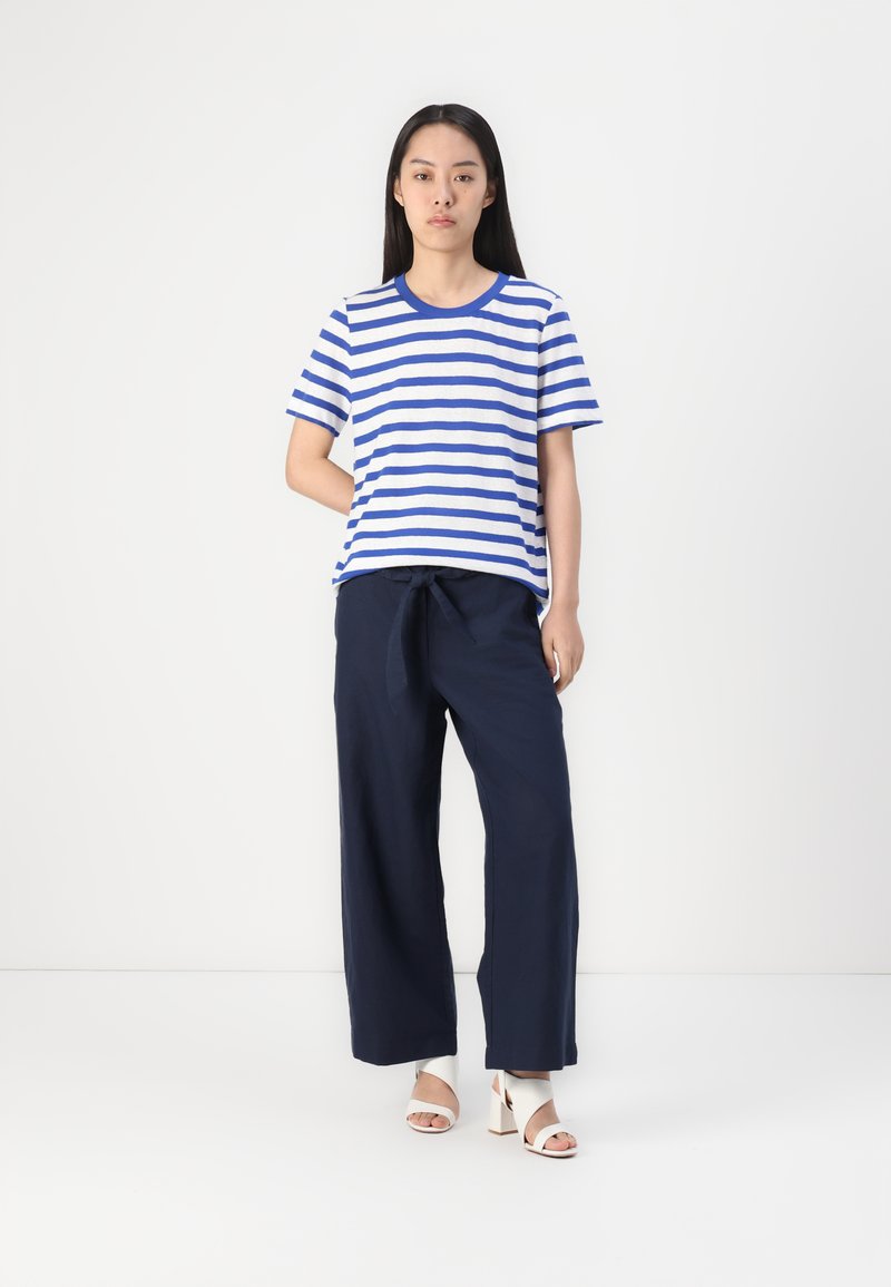 Gestreiftes Baumwoll-T-Shirt in Blau und Weiß, kombiniert mit engen, navyfarbenen Hosen mit weitem Bein und gebundenem Bund. Das Modell trägt weiße Blockabsatzsandalen.