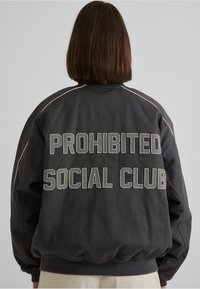Jachetă de tip bomber neagră, cu textul "PROHIBITED SOCIAL CLUB" brodat pe spate. Manșetele și tivul ribbate, cu detalii de coasere în contrast.