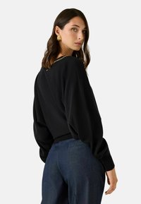 Sort sweater med en afslappet pasform, ballonærmer og en kædeaccent bagpå. Kombineret med mørkeblå bukser. Glat tekstur.