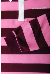 Chemise à manches longues avec des rayures horizontales roses et violet foncé, col blanc et texte brodé « COLOURFUL REBEL » près du col.