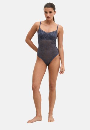 Femme debout pieds nus portant un body bleu marine transparent à fines bretelles avec un motif ondulé subtil sur un fond clair uni.